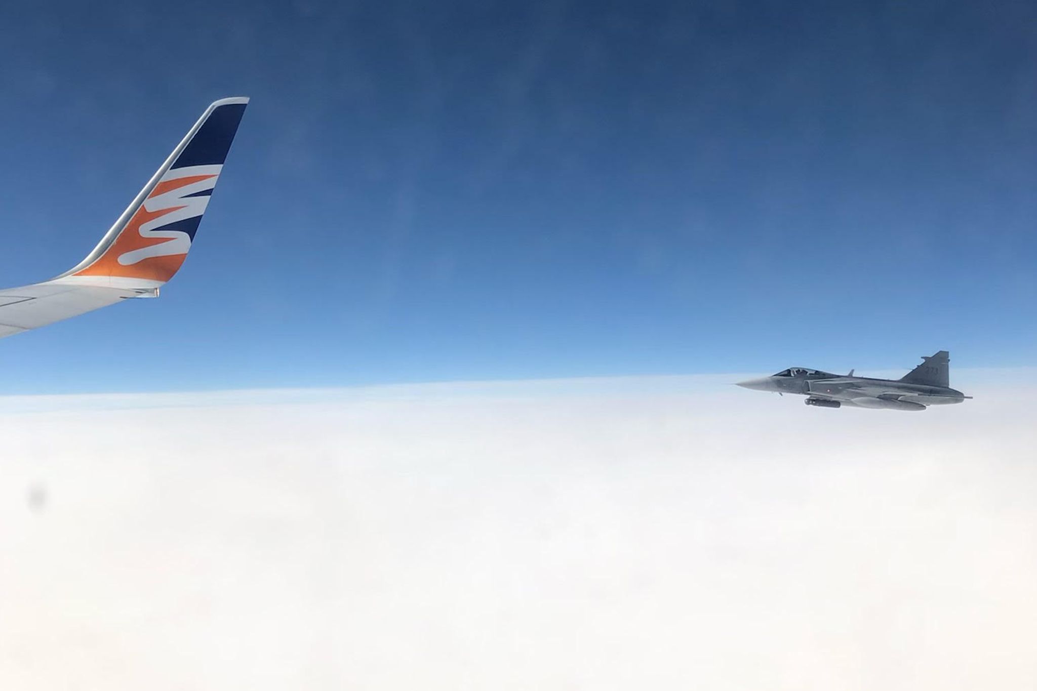 Från flygplansfönstret uppe bland molnen där man ser vingspetsen på flygplanet och en Jas 39 Gripen flyga jämsides och eskortera planet.